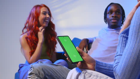 Close-Up-Of-Woman-With-Green-Screen-Mobile-Phone-Sitting-With-Gen-Z-Friends-Talking-And-Sharing-Posts-3
